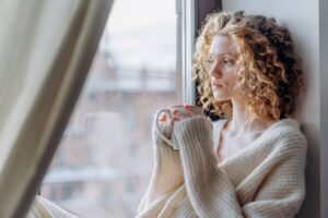 Frau sitzt mit einer Tasse am Fenster und blickt nachdenklich nach draußen